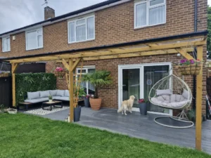 Pergola adosada de madera clara sobre terraza de plataforma gris con sofa rinconero silla colgante tipo nido y macetas con flores