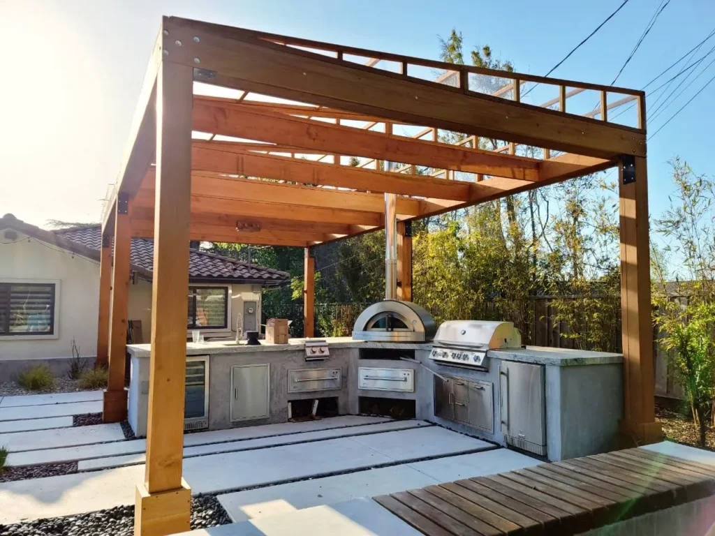 Cocina exterior modular bajo pérgola de madera con horno de leña, parrilla de acero inoxidable y encimeras de hormigón pulido . Cocina exterior modular bajo pergola de madera con horno de lena parrilla de acero inoxidable y encimeras de hormigon pulido