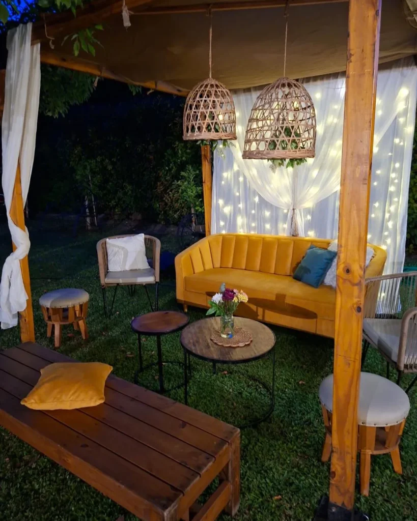 Pérgola de jardín con sofá mostaza, cortinas blancas con luces integradas, lámparas de mimbre y mesas circulares de madera Pergola de jardin con sofa mostaza cortinas blancas con luces integradas lamparas de mimbre y mesas circulares de madera